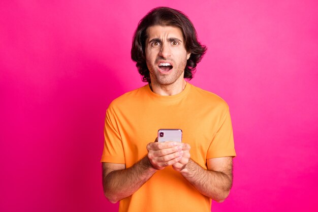 Foto eines jungen depressiven Kerls, der das Telefon mit offenem Mund hält, trägt orangefarbenes T-Shirt isoliert rosa Hintergrund