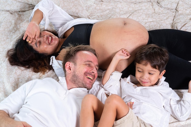 Foto eines jungen, fröhlichen, positiv lächelnden Paares, schwangere Frau und Ehemann mit ihrem Sohn, der sich im Bett entspannt