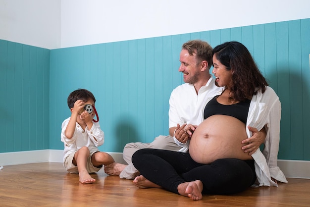 Foto eines jungen, fröhlichen, positiv lächelnden Paares, schwangere Frau und Ehemann mit ihrem Sohn, der sich zu Hause entspannt