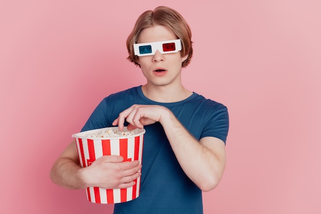 Foto eines jungen mannes, der popcorn-papierbox hält und eine 3d-brille trägt, die einen isolierten rosafarbenen hintergrund ansieht