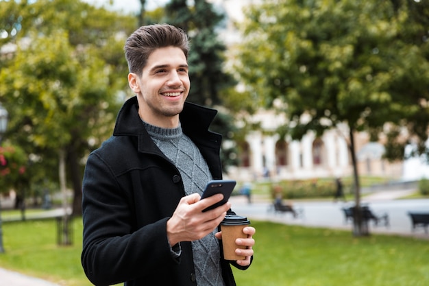 Foto eines kaukasischen Mannes der 30er Jahre, der eine Jacke trägt, die Smartphone hält und Kaffee zum Mitnehmen trinkt, während er durch den Stadtpark geht