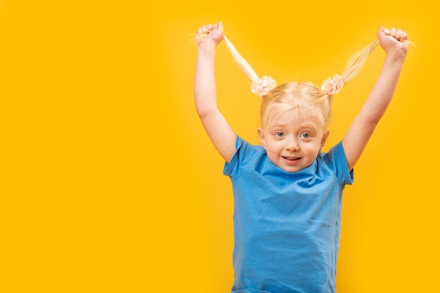 Foto eines lustigen kleinen Mädchens in blauem T-Shirt, das Spaß hat und zwei Pferdeschwänze mit ihrem blonden Haar hält Gelber Hintergrund Kopierbereich