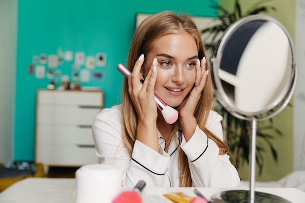 Foto eines positiv optimistischen lächelnden jungen süßen Mädchens im Pyjama zu Hause in der Nähe des Spiegels mit Make-up-Pinsel.