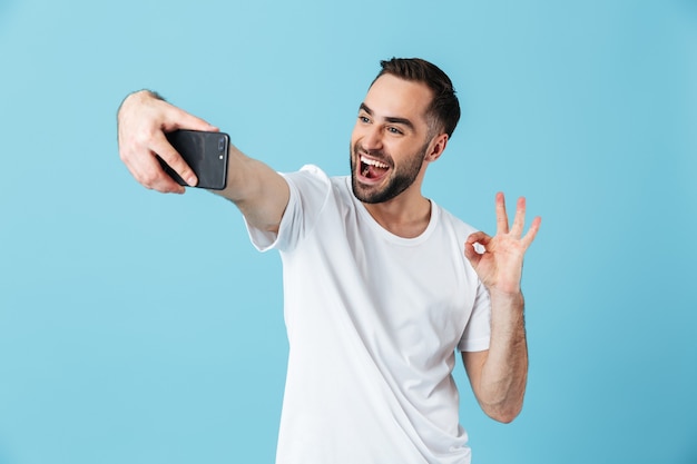 Foto eines positiven brünetten Mannes, der ein einfaches T-Shirt trägt, lacht und ein Selfie auf dem Smartphone macht, isoliert über blau