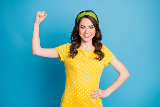 Foto eines positiven fröhlichen Mädchens, das ihre Handmuskeln zeigt, die ein Polka-Dot-Outfit tragen, das auf blauem Hintergrund isoliert ist?