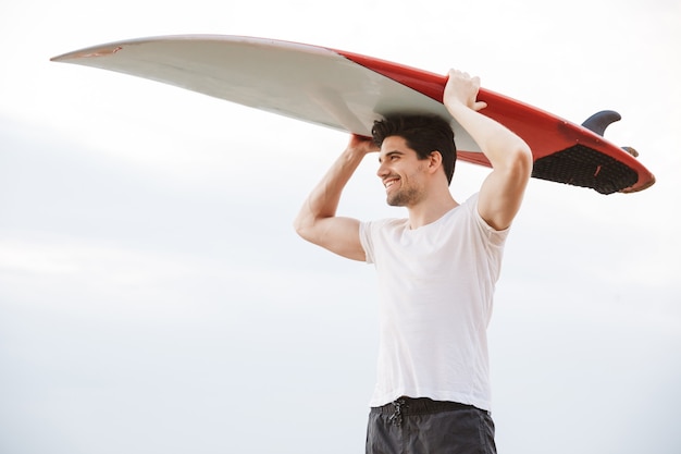 Foto eines positiven glücklichen gutaussehenden Mannsurfers mit dem Surfen an einem Strand draußen.