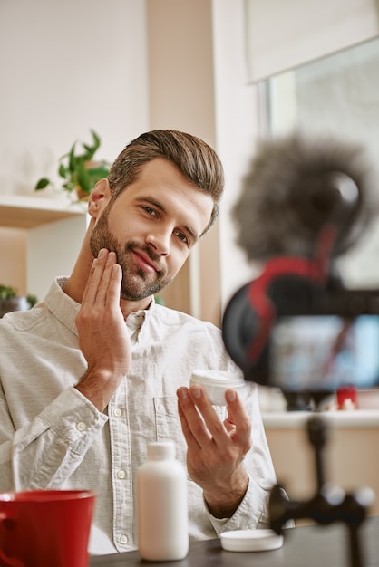 Foto eines positiven männlichen Beauty-Bloggers, der während des Live-Streamings kosmetische Creme auf sein Gesicht aufträgt.