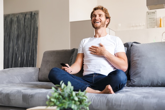 Foto eines schönen glücklichen jungen Mannes auf Sofa zu Hause, das ferngesteuertes Fernsehen fernhält.