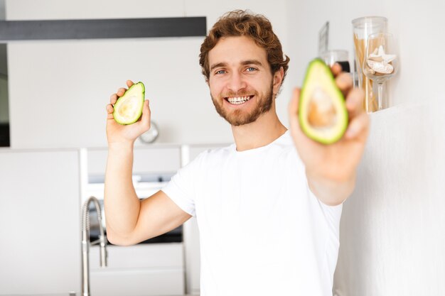 Foto eines schönen jungen Mannes an der Küche zu Hause, die Avocado kochend hält.