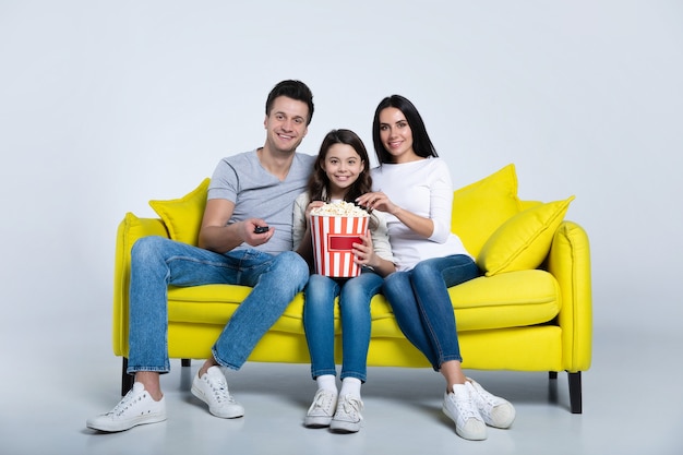 Foto eines süßen Kindes und seiner Eltern, die zusammen Popcorn essen, während sie auf der Couch Fernsehsendungen ansehen.