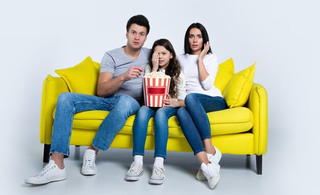 Foto eines süßen Kindes und seiner Eltern, die zusammen Popcorn essen, während sie auf der Couch Fernsehsendungen ansehen.