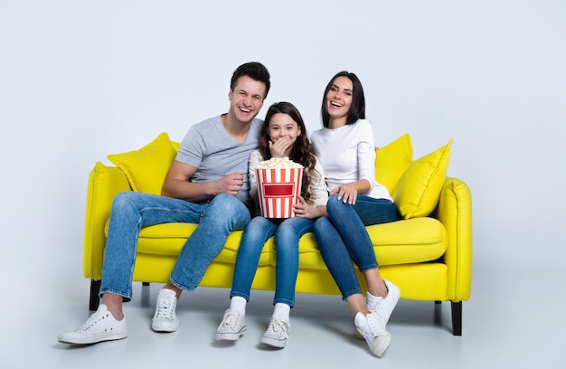 Foto eines süßen Kindes und seiner Eltern, die zusammen Popcorn essen, während sie auf der Couch Fernsehsendungen ansehen.