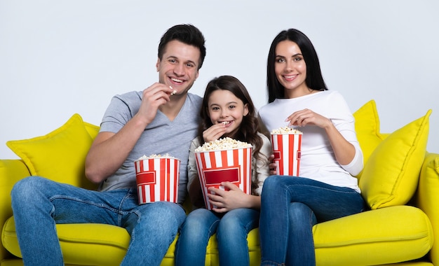 Foto eines süßen Kindes und seiner Eltern, die zusammen Popcorn essen, während sie auf der Couch Fernsehsendungen ansehen.