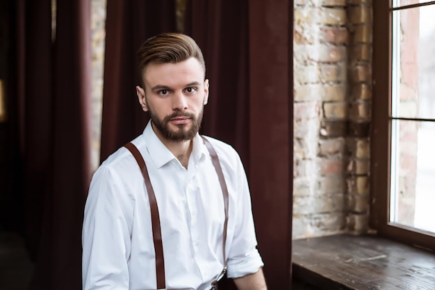 Foto elegante e elegante de um cara barbudo bonito em camisa branca, sentado na cadeira no estúdio