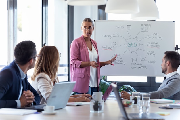 Foto de elegante joven empresaria apuntando a la pizarra blanca y explicar un proyecto a sus colegas en el lugar de coworking.