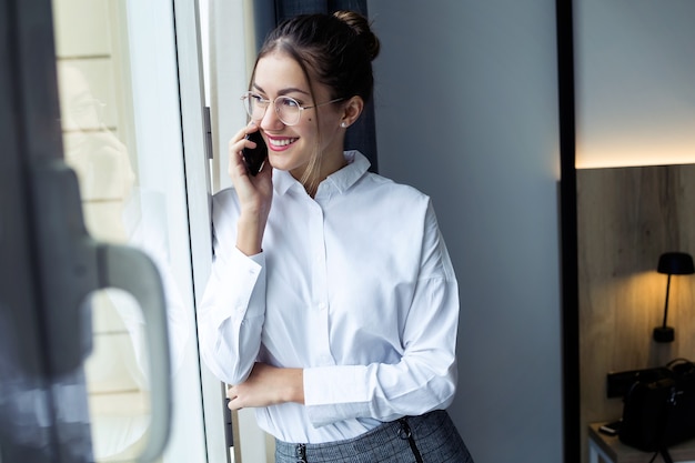 Foto de elegante joven empresaria hablando con su teléfono inteligente y mirando hacia los lados de pie cerca de la ventana en la habitación del hotel.