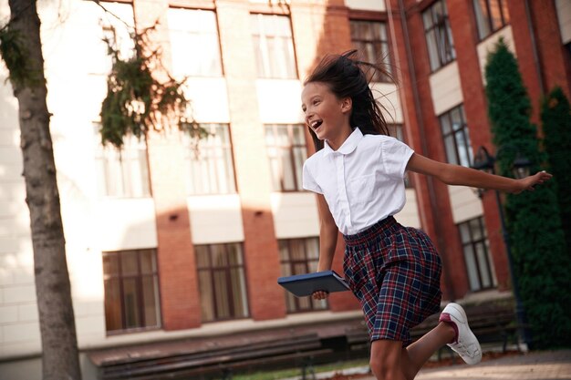 Foto em ação correndo Menina de escola em uniforme está ao ar livre perto do edifício