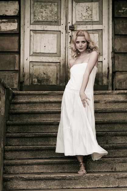 Foto em preto e branco de uma bela jovem em um vestido branco desce as escadas no fundo de um antigo prédio com portas. Conceito de romance gótico