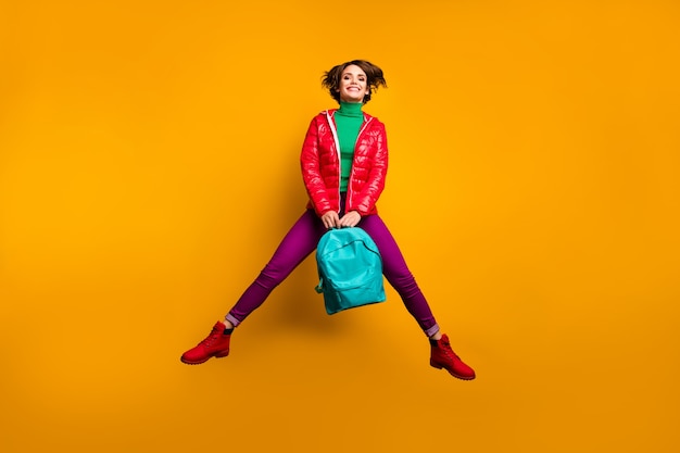 Foto em tamanho grande de uma garota estudantil jovem alegre, divertida e divertida, pular, segurar, mochila azul, desfrutar das emoções, usar calças verdes roxas, calças, botas vermelhas