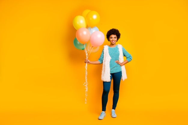 Foto em tamanho real de uma garota afro-americana alegre segurando balões de ar, ela está no aniversário, usando roupa branca azul fofa e sapatos elegantes da moda isolados sobre a parede de cor amarela