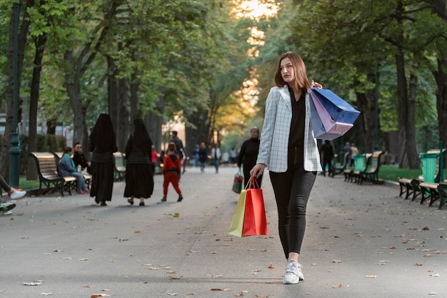 Foto em tamanho real de uma garota de jaqueta com sacolas de compras de papel caminhando pelo parque Compras bem-sucedidas Vendas com desconto