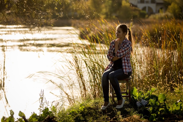Foto em tons de uma jovem sentada ao pôr do sol à beira do lago
