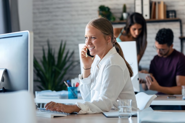 Foto de empresaria inteligente hablando por teléfono móvil mientras trabaja con su computadora en la oficina.