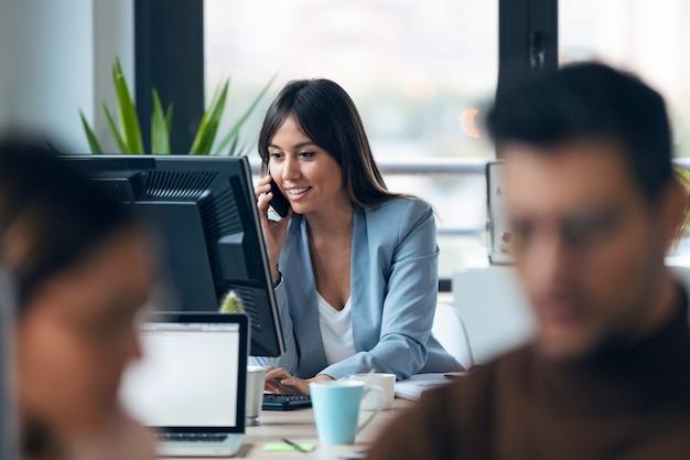 Foto de empresaria joven inteligente hablando con el teléfono móvil mientras trabaja con la computadora en el espacio de trabajo moderno.