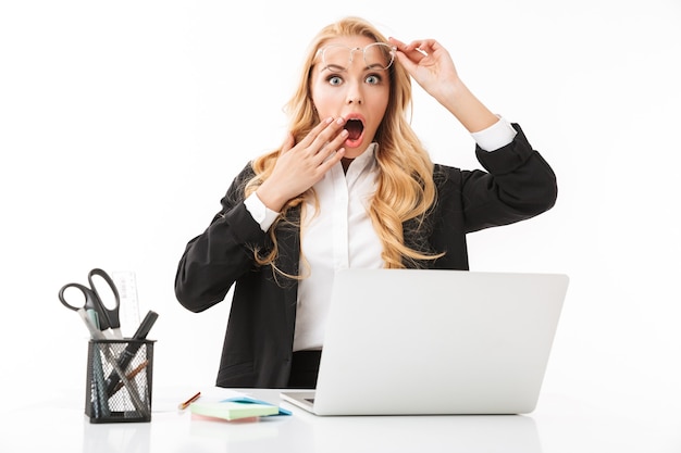 Foto de empresaria sorprendida sentada en el lugar de trabajo y trabajando en la computadora portátil, aislado