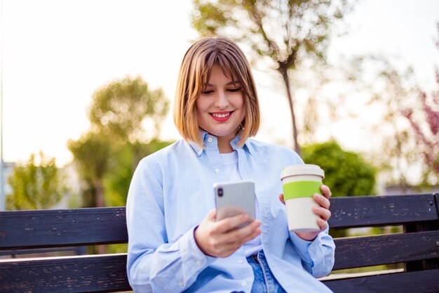 Foto de una encantadora y bonita dama que se comunica con un moderno banco para sentarse sonriendo afuera