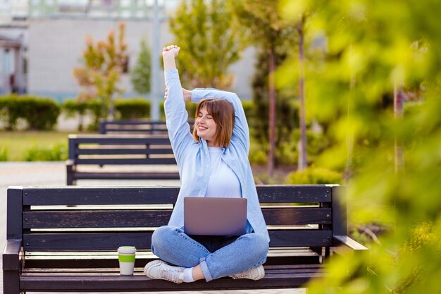 Foto de una encantadora chica cansada de cabello castaño levantando las manos estirando la columna trabajando en línea en una laptop