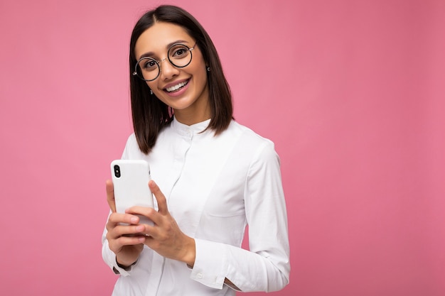 Foto de encantadora mujer morena sonriente joven vistiendo blusa blanca y gafas ópticas de pie