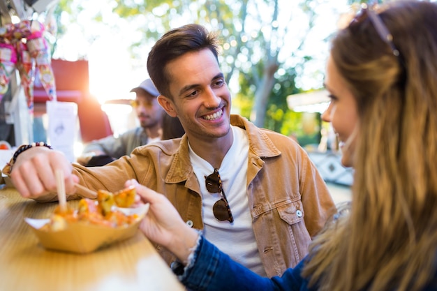 Foto de la encantadora pareja atractiva comiendo patatas juntos en el mercado de comer en la calle.