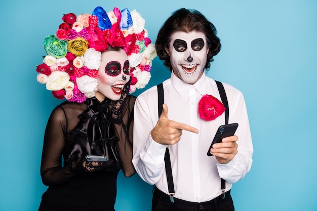 Foto de encantadora pareja espeluznante hombre dama sostener directo dedo teléfono encontrar amigo no muerto red social vestir vestido negro muerte disfraz rosas diadema tirantes aislado color azul fondo