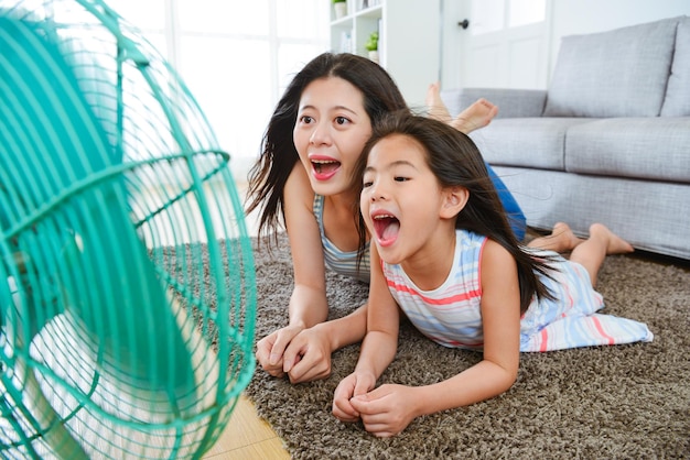 foto de enfoque selectivo de una mujer dulce y una niñita feliz jugando a un ventilador eléctrico y soplando viento fresco en casa para eliminar el calor del verano.