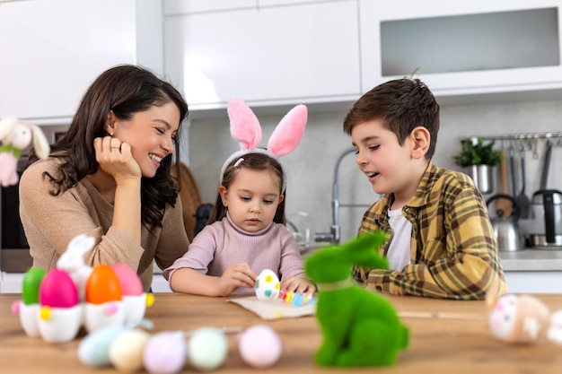 Foto foto engraçada de uma família alegre, feliz, mãe, filho e filha se divertindo e brincando enquanto decoram ovos de páscoa juntos para as férias de primavera sentados à mesa em uma cozinha aconchegante