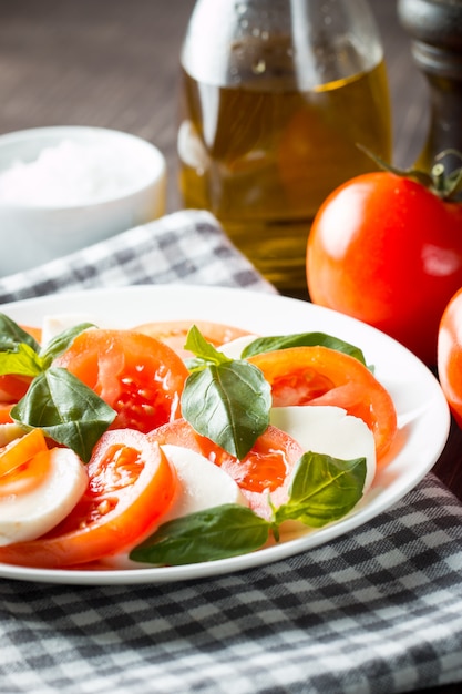 Foto de ensalada Caprese con tomate, albahaca, mozzarella, aceitunas y aceite de oliva sobre fondo de madera. Ingredientes de ensalada caprese tradicional italiana. Concepto de comida mediterránea, orgánica y natural.