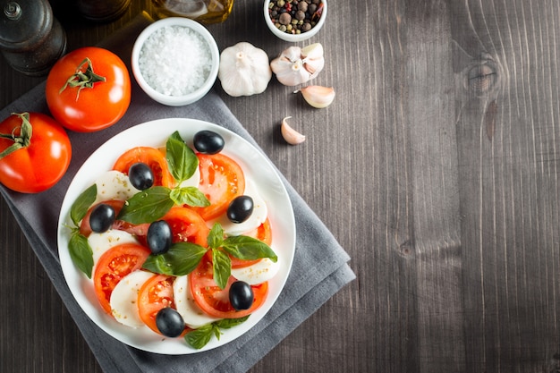 Foto de ensalada Caprese con tomate, albahaca, mozzarella, aceitunas y aceite de oliva sobre fondo de madera. Ingredientes de ensalada caprese tradicional italiana. Concepto de comida mediterránea, orgánica y natural.