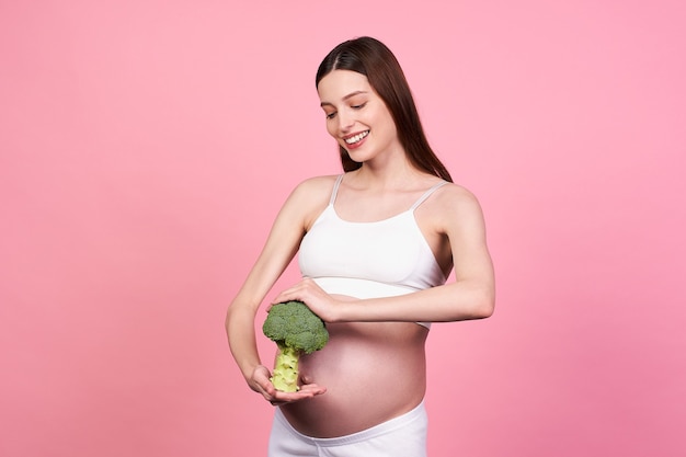 Foto de una esbelta chica embarazada bastante blanca sosteniendo brócoli en sus brazos y posando con el vientre desnudo, mirando con una sonrisa, futura madre en ropa deportiva. Concepto de embarazo y nutrición.