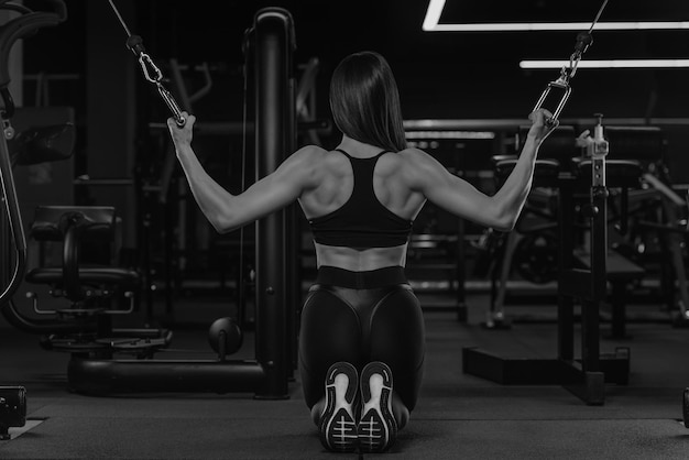 Una foto de espaldas de la mujer deportiva que está haciendo empuje vertical en el cruce de la máquina de cable de ambos bloques de rodillas. Una chica musculosa usa un top y pantalones cortos elásticos de cintura alta en un gimnasio.