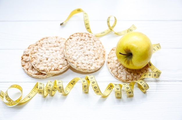 Foto esportiva Estilo de vida saudável Conceito de fitness Pão e frutas crocantes com baixo teor de gordura Fita métrica para corpo em forma Fundo de madeira branca Configuração plana perfeita