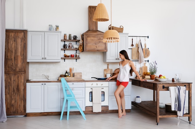 Foto foto de estilo de vida de linda chica de pie junto a la estufa en la cocina, cocinando y oliendo agradables aromas de freír alimentos