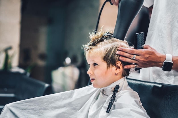 Foto de estilo de vida Peluquería rubio niño cliente Peluquero hombre trabajo mover mano peine herramienta tijeras secador clip capa blanca Estilo moderno niño corte de pelo corto Servicio de negocios de moda para niños