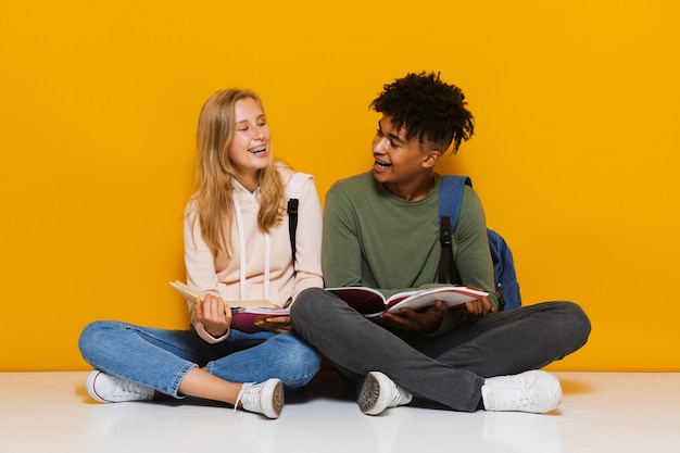 Foto de estudiantes adolescentes de 16 a 18 años que usan libros de lectura mientras están sentados en el piso con las piernas cruzadas, aislado sobre fondo amarillo
