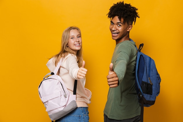 Foto de estudiantes felices, hombre y mujer de 16-18 con mochilas riendo y mostrando los pulgares hacia arriba, aislado sobre fondo amarillo