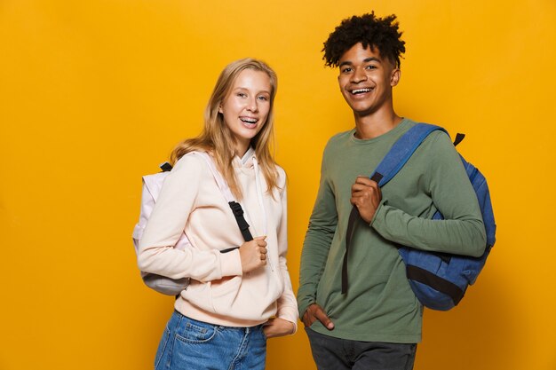 Foto de estudiantes felices, hombre y mujer de 16-18 con mochilas riéndose de la cámara, aislado sobre fondo amarillo