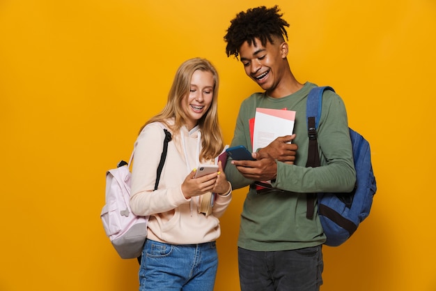 Foto de estudiantes universitarios hombre y mujer de 16-18 con mochilas sonriendo mientras sostiene libros de ejercicios y teléfono móvil, aislado sobre fondo amarillo