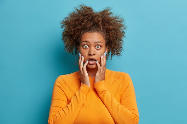 Foto de estudio de aterrorizada niña adolescente de pelo rizado agarra la cara tiene ojos saltó miradas con incredulidad