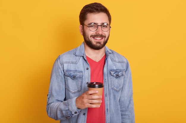 Foto de estudio de atractivo joven con barba y cabello oscuro, feliz expresión facial, sosteniendo una taza de café o té de papel, vestidos elegantes chaqueta vaquera y camisa roja