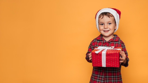 Foto de estudio de un bebé en pijama de Navidad y un sombrero sobre un fondo amarillo con una caja de regalo roja brillante en sus manos Un lugar para su publicidad de texto